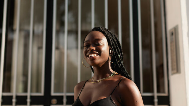 A Young Black Woman Smiles Happily Looking At The Camera With The Front Door Of Her House Behind Her