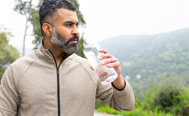 Bearded ethnic sportsman standing with drinking water bottle in daytime