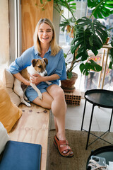 Beautiful smiling blond woman in hipster cafe holding her anxious dog Jack Russell terrier. Sunny...