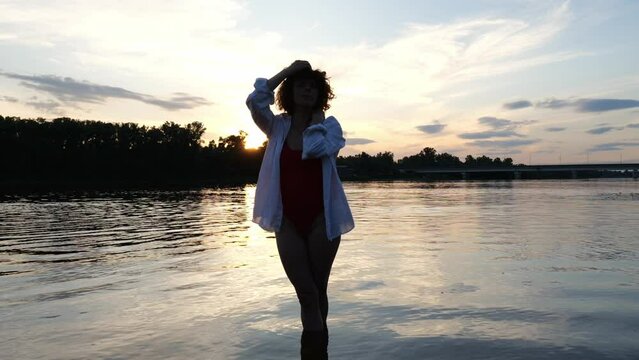 A young woman in her thirties in a red swimsuit and white shirt poses in dark water at sunset in the evening. Slow motion woman swimsuit relax