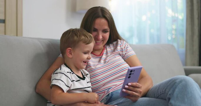 Woman and her little son sitting on cozy sofa with smartphone