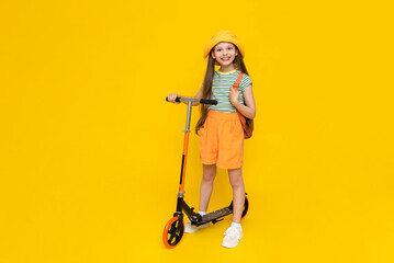A full-length young girl in a hat and shorts is standing next to a scooter and enjoying a summer...