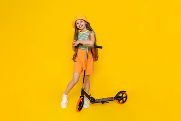 Fototapeta na wymiar A full-length young girl in a hat and shorts is standing next to a scooter and enjoying a summer vacation. Active city recreation for children. Yellow isolated background.
