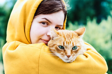 Girl hugging a cat close-up. A beautiful young girl is holding a cute ginger cat on a blurred background. Portrait of a young hipster woman hugging her good friend ginger cat. Best friends.