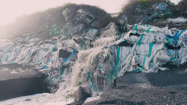 Beautiful slow motion view of dirty river streaming on cascade of filthy plastic sacks with sand and rubbish. Water pollution. Chemicals in drain river. Industrial refuse waste. Ecological problems.
