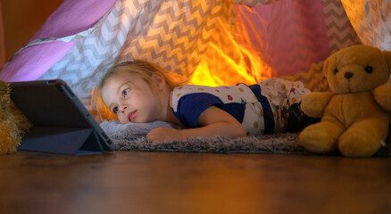 Cute little caucasian girl kid watching video on digital tablet enjoying online internet entertainment, free play time relaxing sleeping in a tent comfortable, childhood technology video game
