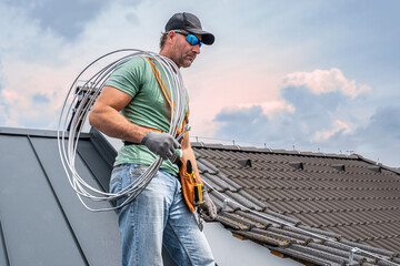 Work on the roof. An electrician installing a lightning rod conductor on a tiles roof. - obrazy, fototapety, plakaty
