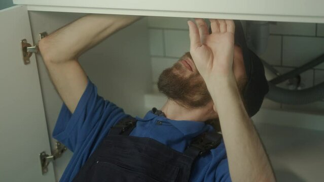 Young Plumber with Long Hair, Beard, and Hat Repairs Sink