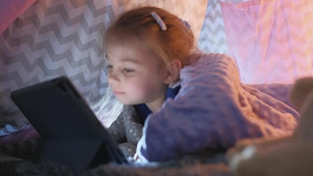 Cute little caucasian girl kid watching video on digital tablet enjoying online internet entertainment, free play time relaxing sleeping in a tent comfortable, childhood technology video game