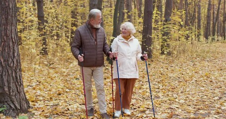 Two sporty active happy old pensioners enjoy nordic walk in nature wood park in autumn. Senior elder couple stroll Romantic family hikers date. Elderly grandparents training in the forest Slow motion - Powered by Adobe