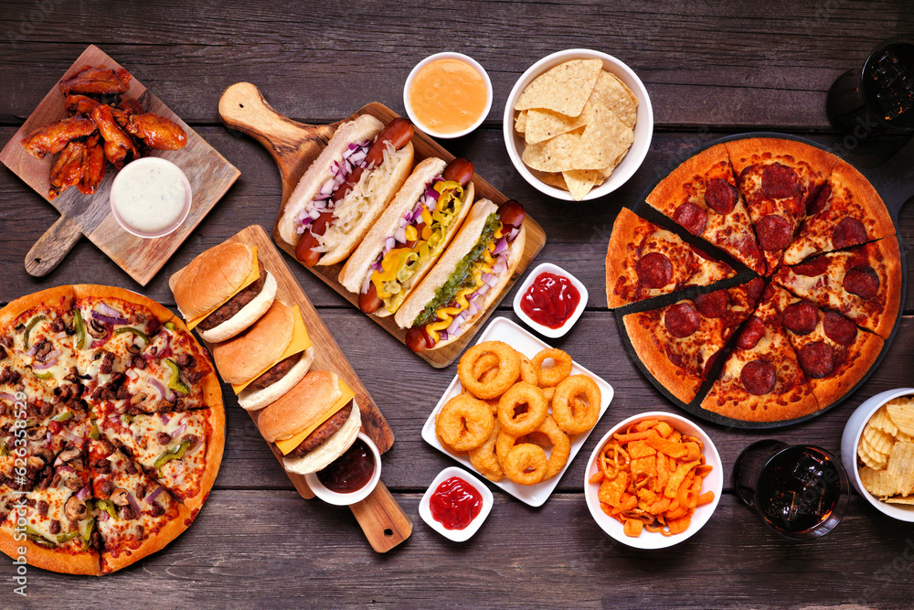 Wall mural Junk food table scene. Pizza, hamburgers, chicken wings, hot dogs and salty snacks. Above view over a dark wood background.