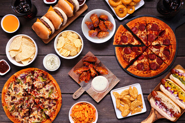 Junk food table scene. Pizza, hamburgers, hot dogs, chicken wings and salty snacks. Overhead view over a dark wood background. - obrazy, fototapety, plakaty