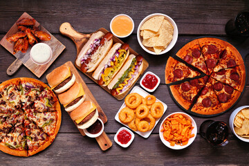 Junk food table scene. Pizza, hamburgers, chicken wings, hot dogs and salty snacks. Above view over a dark wood background.
