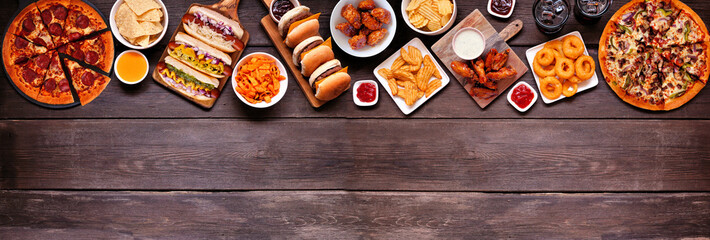 Junk food top border. Pizza, hot dogs, hamburgers, chicken wings and salty snacks. Above view over a dark wood banner background with copy space.