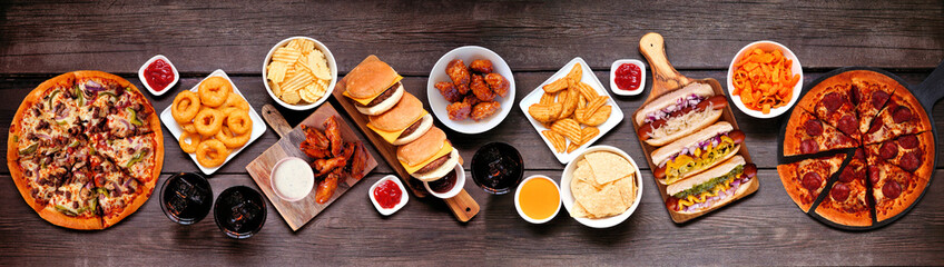 Junk food table scene. Pizza, hamburgers, chicken wings, hot dogs and salty snacks. Top down view...