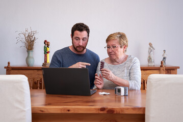 Senior woman accompanied with her adult son showing a blister pill pack and listening to her doctor in a online medical consultation at home.