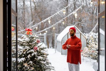 Mann decorates Christmas tree with festive ballls while stands on terrace at snowy backyard. Girl preparing for a winter holidays