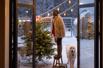 Young woman decorates Christmas tree, attaching festive bow on top while standing on step ladder at...