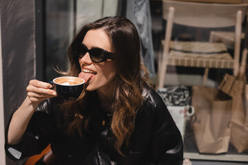 Portrait of laughing blonde girl with coffee in coffee shop terrace. Lovable lady sitting near window and holding cup of coffee and look happy. Woman drink, hold coffee mug, girl lick cup cappuccino.