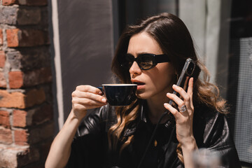 Confused puzzled blonde woman in casual clothes, sits in cafe outside, talking by smartphone, have call conversation look questioningly drinking cappuccino. Girl in glasses getting surprising bad news