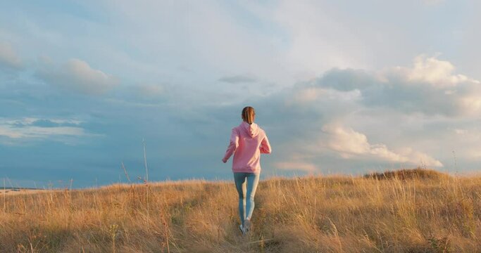 back rare view of fit slim woman in pink sweater and leggins going jogging at agricultural field during summer day slow motion Free spare time Health body care Wellness. fitness, girl burns callories