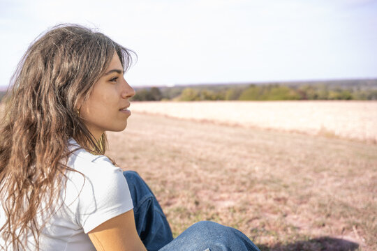 Jeune femme en jean assise au bord d'un champs regardant l'horizon