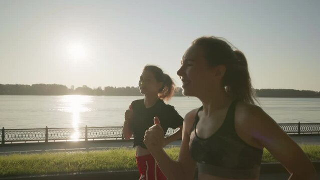 Two beautiful girls jogging at sunset or sunrise on the river embankment. Young multiracial women running together. Brunette and blonde are smiling. Feeling of freedom while running. Concept of cardio
