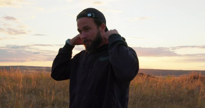 handsome bearded man in cap and hoodie standing on the field in the countryside. Male in sportswear enjoying watching sunset, guy puts on cap, preparing to work out outdoors Slow motion