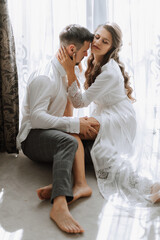 Beautiful, sexy bride in a white robe, groom in a white shirt hugs and kisses the bride against the background of a window in a hotel room. Wedding portrait of newlyweds in love. Vertical photo.