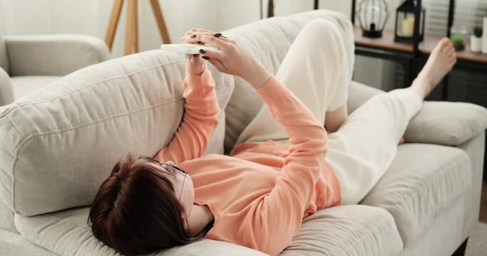 In the living room, a teenage girl lies on the couch, engrossed in a messaging conversation on her phone. Relaxed, she taps away on the screen, exchanging messages with a sense of calm and focus.