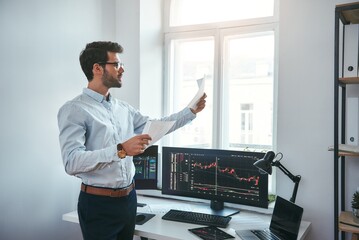 Trading strategy. Smart and young trader in eyeglasses looking at financial reports and analyzing trading charts while standing in front of computer screens in modern office.