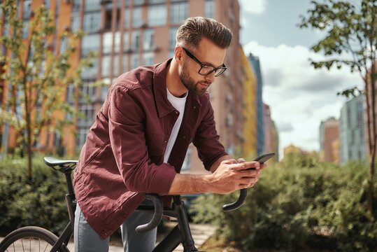 Surfing the net. Side view of handsome man with stubble in casual clothes and eyeglasses leaning at his bicycle and looking at mobile phone while standing outdoors
