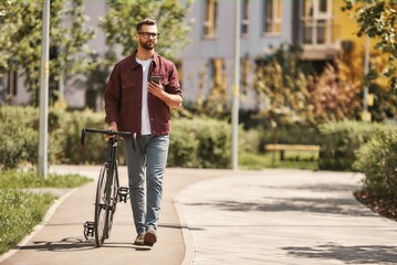My way. Handsome man with stubble in casual clothes and eyeglasses pulling his bicycle and holding smartphone while walking outdoors