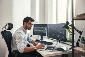 Busy working day. Young bearded trader in eyeglasses working with laptop while sitting in his modern office in front of computer screens with trading charts.