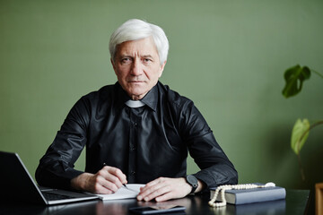 Front view portrait of white haired senior priest working at desk and looking at camera in office...
