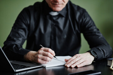 Closeup of senior priest writing in notebook at desk while working on speech in office, copy space