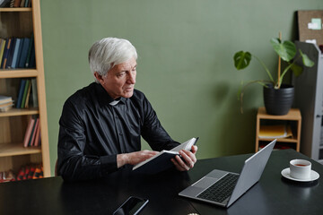 Portrait of white haired senior priest reading Bible at desk in office against green wall, copy space