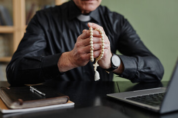 Closeup of senior priest holding rosary while praying at desk in office, copy space