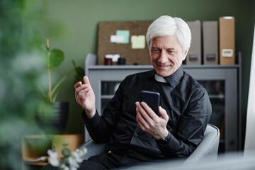 Portrait of smiling senior priest using smartphone and talking by video chat in office, copy space - obrazy, fototapety, plakaty