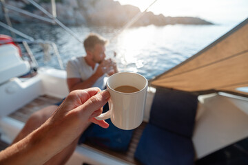 hand with a cup of coffee in the foreground, while in the background a man on a yacht anchored in...