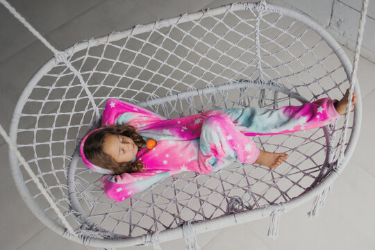 Happy little girl on the porch lying on a wicker swing and playing in pink pajamas with her little toy