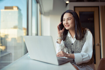 Young smiling Asian business woman client manager working in modern office making phone call. Happy female professional worker talking on mobile cellphone using laptop working at desk.