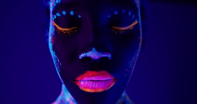 close up face in Neon UF Light. Model Girl with Fluorescent Creative Psychedelic MakeUp Art Design of Female Disco Dancer Model in UV Colorful Abstract Make-Up Slow motion Beauty industry, cosmetology