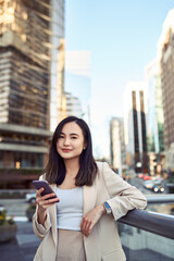 Fototapeta premium Young pretty Asian business woman office professional with cellphone in hands standing on big city urban street holding mobile cell phone, using digital apps on smartphone device, vertical.