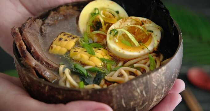 Person holding a ramen bowl in hands, Asian beef soup served with meat slices, topped with boiled egg, noodles soup with udon and seaweed, close up detailed video, 4k footage