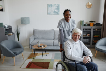 Portrait of white haired senior man in wheelchair smiling at camera indoors with young female nurse assisting, copy space