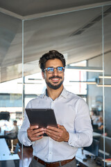 Smiling busy young Latin business man entrepreneur using tablet standing in office at work. Happy...