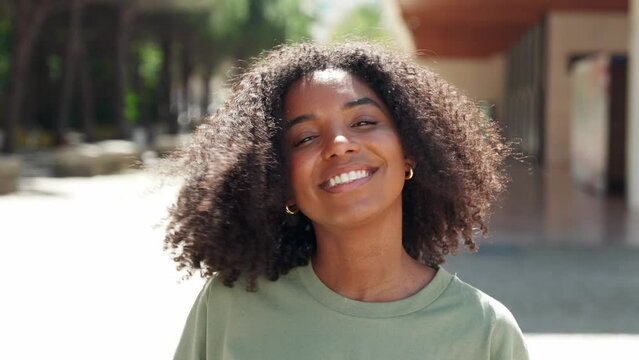Young happy cute pretty young beautiful African American ethnic woman model playing with curly hair looking at camera in city street outdoors on sunny day, close up face portrait.