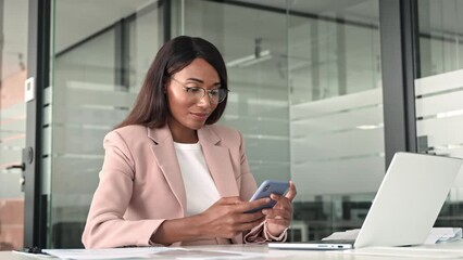 Young busy professional African American business woman executive manager holding cellphone, using mobile device looking at cell phone technology sitting at desk in office working on laptop. - Powered by Adobe
