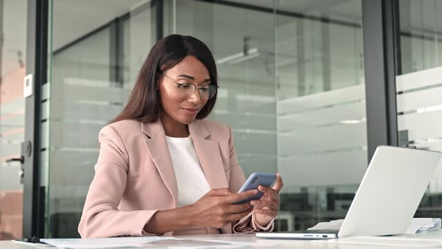 Young busy professional African American business woman executive manager holding cellphone, using mobile device looking at cell phone technology sitting at desk in office working on laptop. - Powered by Adobe